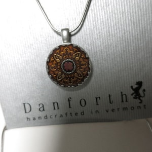 Puede incluir: Un collar de cadena de plata con un colgante redondo que presenta un diseño de mandala dorado y marrón con un centro rojo. El colgante está unido a una cadena de plata. El collar se muestra en una tarjeta blanca con el texto "Danforth handcrafted in Vermont" y un logotipo de león.