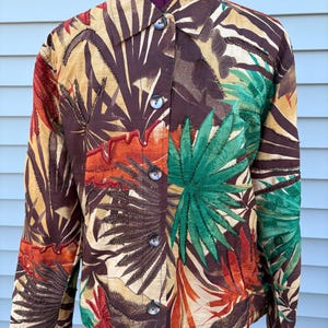 May include: A button-down jacket with a tropical leaf pattern in shades of brown, orange, green, and tan. The jacket has long sleeves and a collared neckline. The fabric appears to be a woven material.