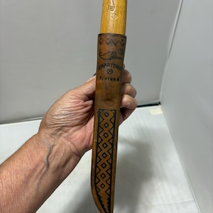 May include: A light brown wooden knife handle and a matching leather sheath with a dark brown geometric pattern. The sheath features the text J. MARTTIINI and Finland, along with a fish graphic and a registered trademark symbol on the front.