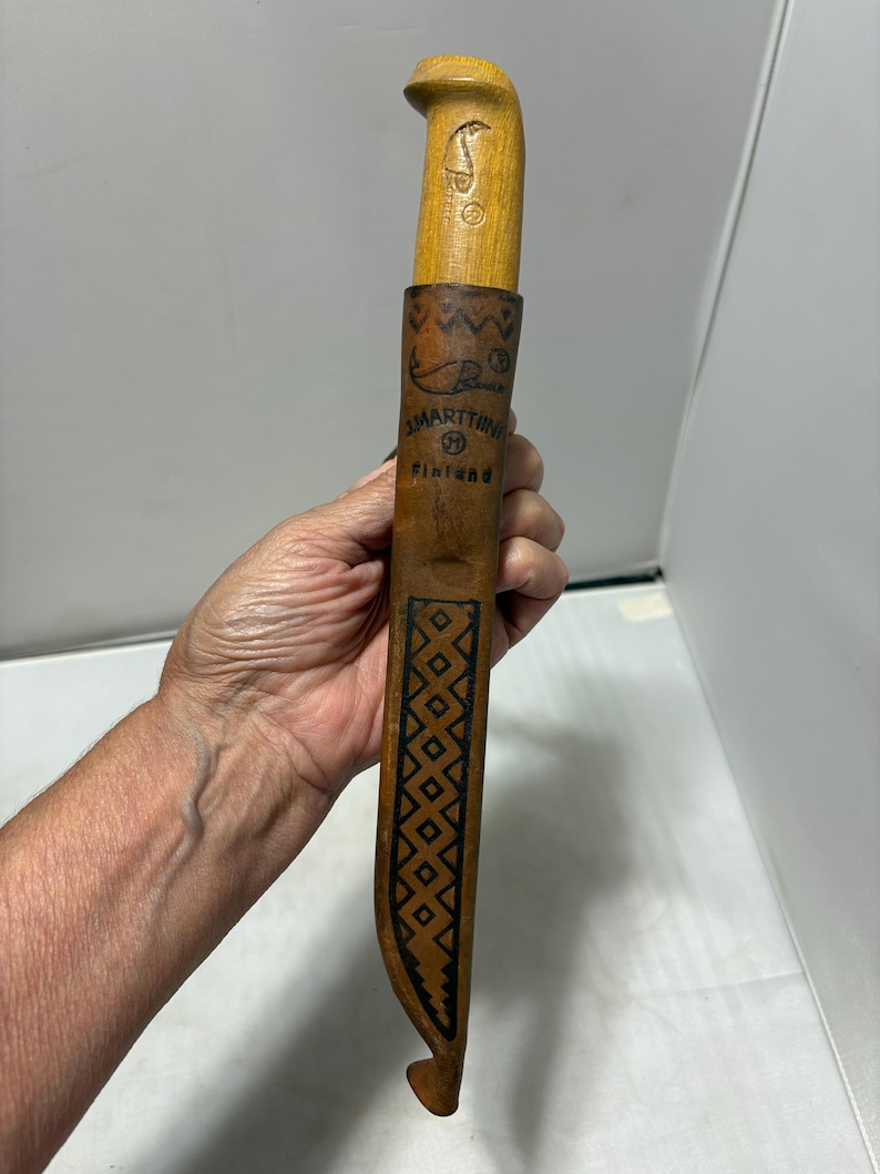 May include: A light brown wooden knife handle and a matching leather sheath with a dark brown geometric pattern. The sheath features the text J. MARTTIINI and Finland, along with a fish graphic and a registered trademark symbol on the front.