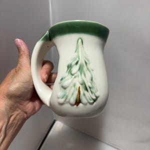May include: A cream-colored ceramic mug featuring a raised, embossed pine tree design in shades of green and brown. The mug has a wide, curved handle and a thick, dark green band around the top rim, creating a distinct contrast against the off-white body.