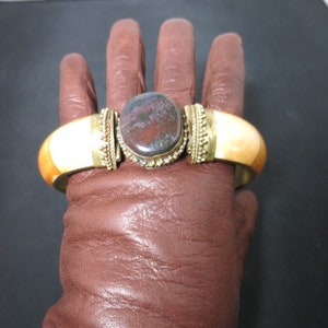 May include: A gold bangle bracelet with a large, oval-shaped, multi-colored stone set in the center. The bracelet is made of bone and has a decorative design.