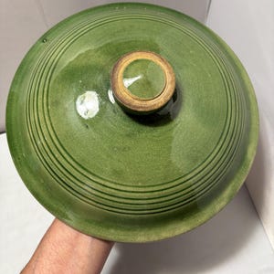 Peut inclure: Un couvercle en céramique vert et rond, orné de stries circulaires concentriques sur toute sa surface. Le couvercle possède un bouton central surélevé avec un bord beige non émaillé. La glaçure verte présente des variations de ton.