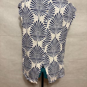 K&ouml;nnte beinhalten: Ein &auml;rmelloses Oberteil mit geteiltem R&uuml;cken, das auf einer Schaufensterpuppe pr&auml;sentiert wird. Das Oberteil hat ein sich wiederholendes Muster aus marineblauen f&auml;cherartigen Formen auf cremefarbenem Hintergrund. Der Stoff scheint ein leichtes Material zu sein.