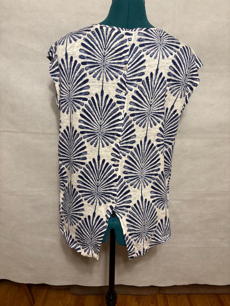 K&ouml;nnte beinhalten: Ein &auml;rmelloses Oberteil mit geteiltem R&uuml;cken, das auf einer Schaufensterpuppe pr&auml;sentiert wird. Das Oberteil hat ein sich wiederholendes Muster aus marineblauen f&auml;cherartigen Formen auf cremefarbenem Hintergrund. Der Stoff scheint ein leichtes Material zu sein.