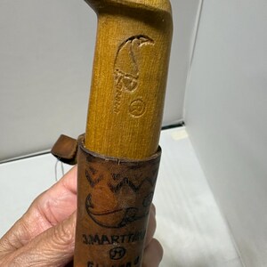 May include: A light brown wooden knife handle with a carved logo and a registered trademark symbol, inserted into a brown leather sheath featuring a stamped geometric pattern, the text J. MARTTIINI, and the word Finland printed on the front.