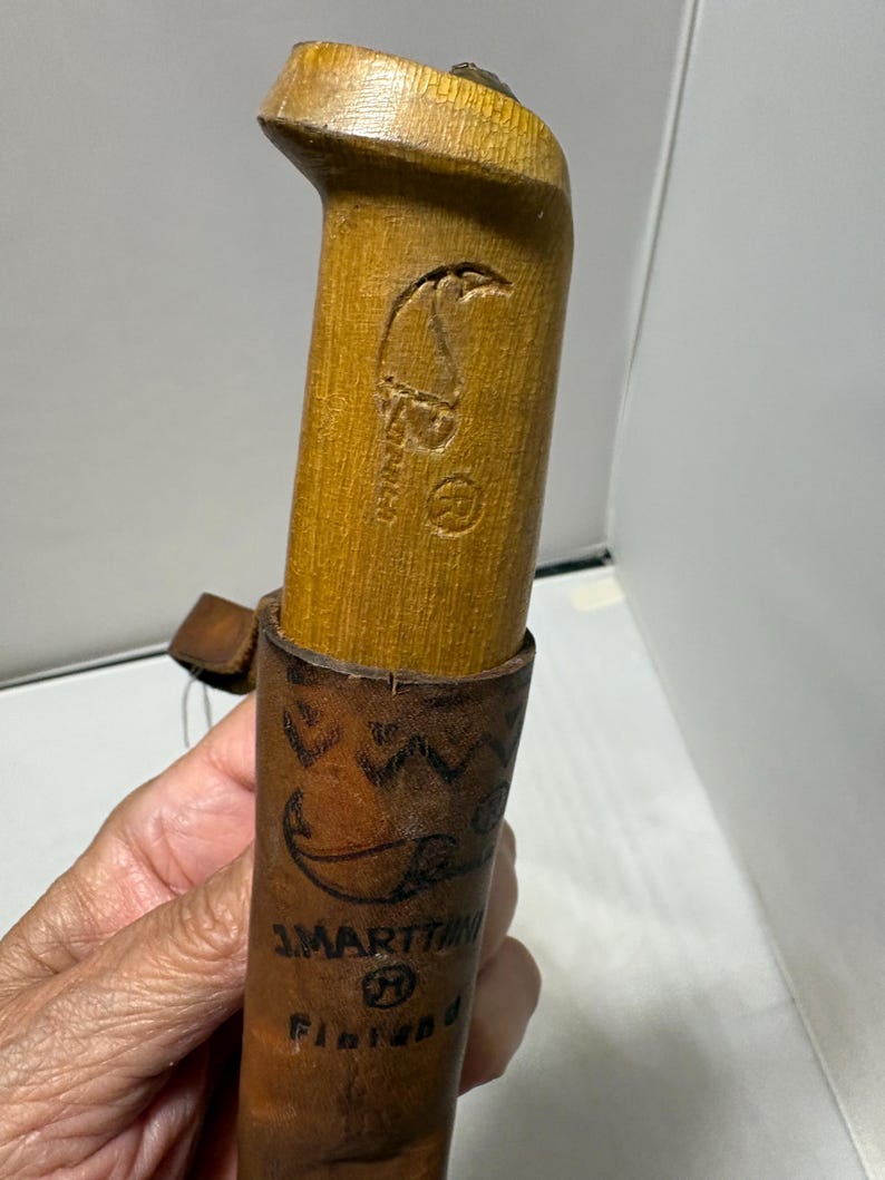 May include: A light brown wooden knife handle with a carved logo and a registered trademark symbol, inserted into a brown leather sheath featuring a stamped geometric pattern, the text J. MARTTIINI, and the word Finland printed on the front.