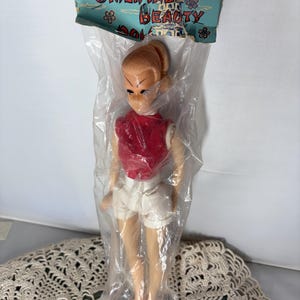 May include: A vintage doll in a plastic bag, wearing a red top, white shorts, and pink shoes. The doll has blonde hair and is labelled "Oriental Beauty" on the packaging. The packaging is light blue with floral and pagoda designs.