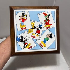 Puede incluir: Una caja cuadrada de madera con una baldosa de cerámica en la parte superior que presenta cinco ilustraciones de Mickey Mouse con forma de pieza de rompecabezas. Las imágenes muestran a Mickey con varios atuendos y los años 1928, 1933, 1940, 1950 y 1988 sobre un fondo azul claro.