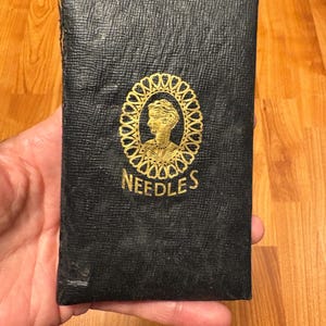 Może przedstawiać: Zabytkowe czarne etui na igły ze złotym wytłoczonym wzorem. Etui posiada portret w owalnej ramie oraz napis "NEEDLES" złotymi literami. Etui jest trzymane w dłoni.