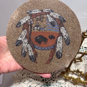 May include: Round, tan-colored decorative plate featuring a Native American design. The design includes a central shield with bison, surrounded by feathers and a dreamcatcher. The plate has a textured surface and a vintage aesthetic.