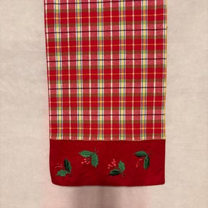 Puede incluir: Un camino de mesa a cuadros rojos y verdes con un borde inferior rojo. El borde presenta hojas de acebo verdes bordadas y bayas rojas. El patrón de cuadros incluye líneas rojas, amarillas, blancas y verdes.