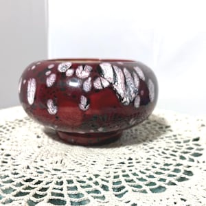 Könnte beinhalten: Eine runde, rote Keramikschale mit glänzendem Finish. Die Schale hat ein gesprenkeltes Muster aus weißen und schwarzen Akzenten. Die Schale steht auf einer weißen Spitzendecke.