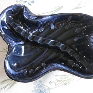 Puede incluir: Un cenicero de cerámica azul con una forma abstracta única. El cenicero tiene un borde elevado texturizado y un acabado liso y brillante.