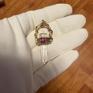 May include: A gold-colored pendant featuring a clear quartz crystal and a rectangular, red gemstone. The pendant has a decorative, woven design around the gemstone and crystal. The pendant is held in a white glove.