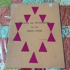 May include: A softcover instructional book titled WEAVES and TECHNIQUES for the INKLE LOOM by Betty Atwood. The cover is light tan with a central diamond pattern formed by eight magenta triangles. A small white price sticker is at the top right.