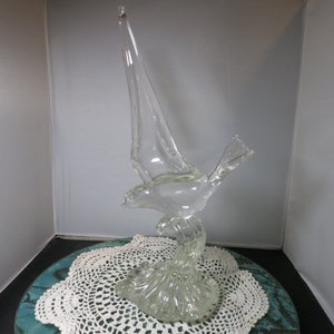 May include: A clear glass sculpture of a bird in flight with its wings spread. The bird is perched on a base that resembles waves. The sculpture is on a white doily.