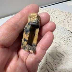 May include: A carved stone figurine of a monkey, featuring tan and black coloration. The sculpture is held in a hand, showcasing the details of the carved features. The monkey figurine is a unique decorative item.