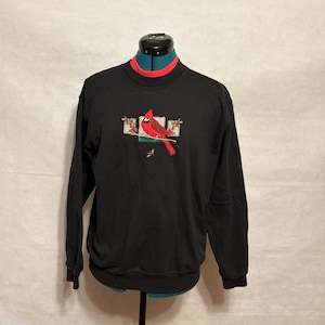 Puede incluir: Sudadera negra con un diseño bordado de un pájaro cardenal rojo y verde. La sudadera tiene un cuello rojo y negro. El pájaro está posado en una rama con bayas rojas. Una cómoda parte superior de manga larga.