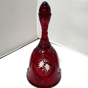 Puede incluir: Una campana de cristal rojo intenso con un mango texturizado y un panel ovalado central decorado con un diseño blanco grabado de dos piñas y agujas. La campana tiene una base acampanada y detalles florales en relieve en el borde inferior.