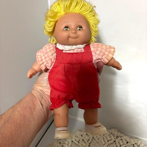 May include: A vintage doll with a smiling face, yellow yarn hair, and blue eyes. The doll wears a pink and white checkered shirt with a white lace collar and red overalls. It has white shoes.