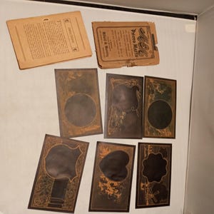 May include: A set of seven vintage photo mats with ornate designs. The mats are made of a dark brown paper with gold accents. The mats are all different shapes and sizes. The mats are labeled "Photo Mats" and "Medal & Memory".