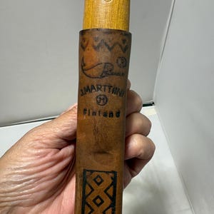 May include: A brown leather sheath for a knife with a wooden handle. The sheath features a stamped fish logo, the text J. MARTTIINI M Finland, and a geometric diamond pattern. The wooden handle above the sheath shows a partial stamped logo.