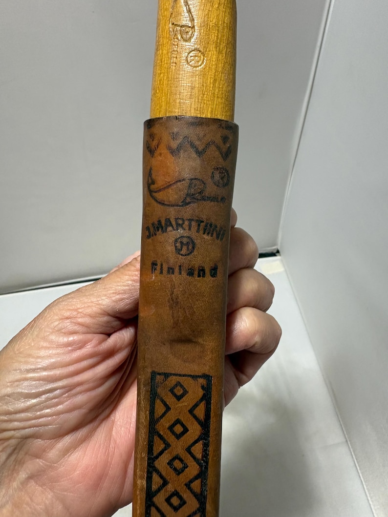 May include: A brown leather sheath for a knife with a wooden handle. The sheath features a stamped fish logo, the text J. MARTTIINI M Finland, and a geometric diamond pattern. The wooden handle above the sheath shows a partial stamped logo.