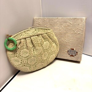 May include: A vintage, light green crocheted clutch with a matching green ring handle. A rectangular box with a striped pattern and a small label is next to the clutch. The clutch has a zipper closure.