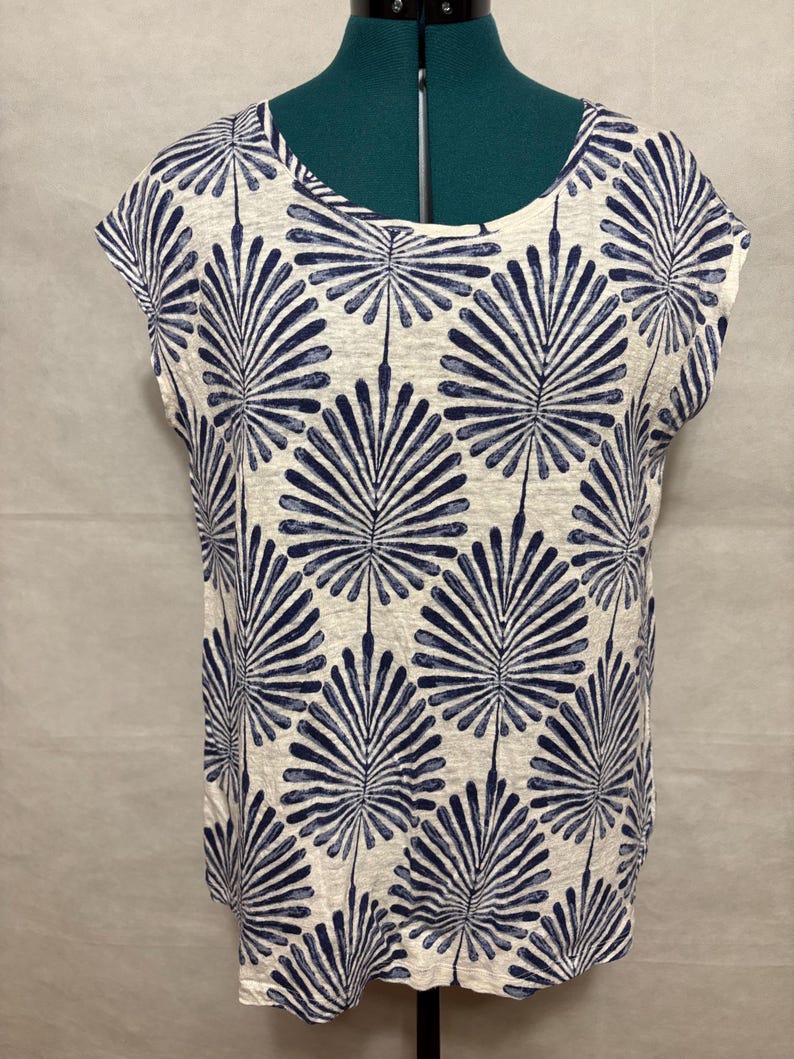 K&ouml;nnte beinhalten: Ein wei&szlig;es und marineblau gemustertes Oberteil mit Rundhalsausschnitt und kurzen &Auml;rmeln. Das Muster zeigt ein sich wiederholendes Design aus stilisierten marineblauen Palmenbl&auml;ttern. Das Oberteil wird auf einer Schaufensterpuppe pr&auml;sentiert.