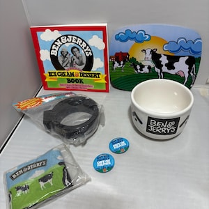 May include: A collection of Ben & Jerry's merchandise, including an ice cream dessert book, a bowl, a cutting board, a black item, a small towel, and two buttons. The items feature the Ben & Jerry's logo and cow imagery.