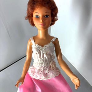 May include: A vintage doll with red hair and blue eyes. The doll is dressed in a white eyelet top and a pink satin skirt. The top has a frilled hem and a small flower. The doll's arms are outstretched.