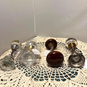 May include: An assortment of vintage door knobs in various styles. The knobs include clear glass, brown, and metal finishes. Some are in a plastic container, and all are displayed on a cream-colored, crocheted surface.