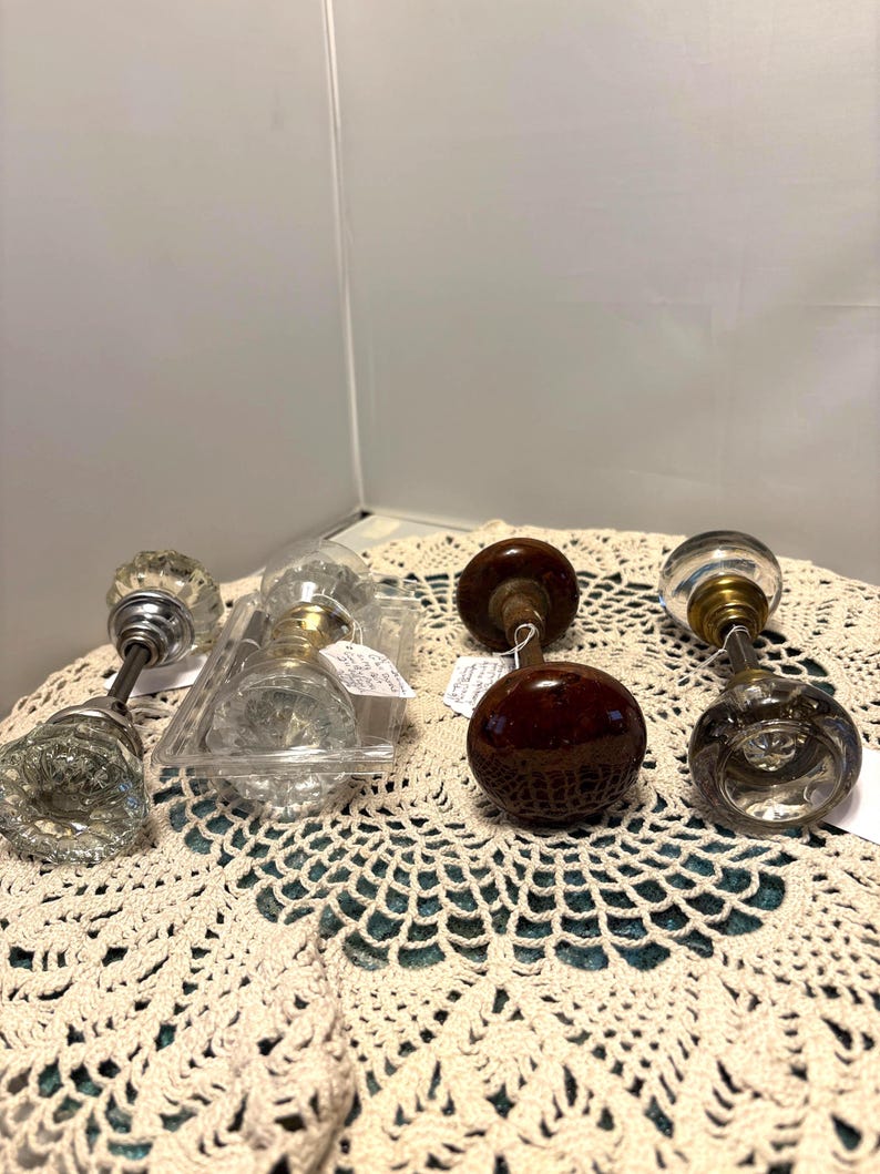 May include: An assortment of vintage door knobs in various styles. The knobs include clear glass, brown, and metal finishes. Some are in a plastic container, and all are displayed on a cream-colored, crocheted surface.