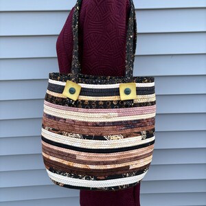 Könnte beinhalten: Handgefertigte Tragetasche mit Steppmuster. Die Tasche hat einen Patchwork aus braunen, schwarzen und cremefarbenen Stoffen. Die Griffe und die obere Zierleiste sind schwarz mit goldenen Akzenten. Zwei quadratische, gelbe Knöpfe befestigen die Griffe an der Tasche.