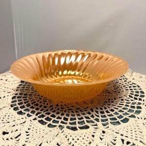 Puede incluir: Un cuenco de vidrio naranja iridiscente con un borde ondulado. El cuenco tiene un interior texturizado y está colocado sobre un tapete de ganchillo crema. El color del cuenco cambia con la luz, creando una estética cálida.