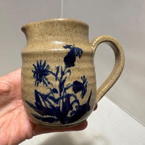 May include: A speckled beige ceramic pitcher featuring a dark blue hand-painted floral design on the front. The pitcher has a rounded body with horizontal ridges, a small pouring spout, and a curved handle on the right side of the vessel.