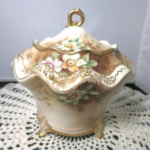May include: A decorative porcelain trinket box with a lid. The box is cream-colored with a brown and gold floral design. The lid has a gold handle. The box has a scalloped edge and sits on three gold feet.