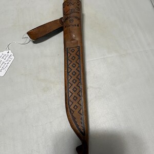 May include: A Marttiini Finland knife with a light brown wooden handle and a tan leather sheath. The sheath features a stamped geometric diamond pattern, a belt loop, and the text Marttiini Finland embossed on the upper front section.