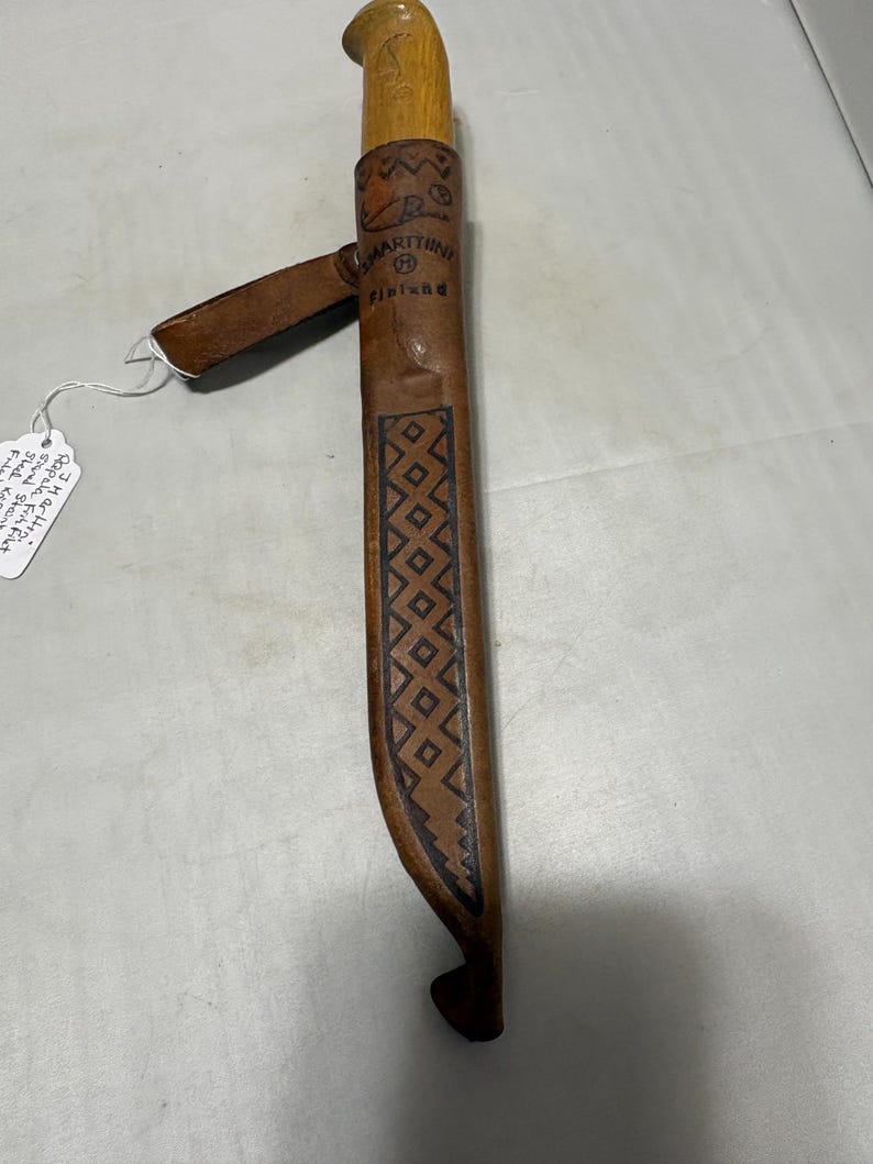 May include: A Marttiini Finland knife with a light brown wooden handle and a tan leather sheath. The sheath features a stamped geometric diamond pattern, a belt loop, and the text Marttiini Finland embossed on the upper front section.