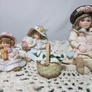 Könnte beinhalten: Drei Porzellanfiguren junger Mädchen. Eine hält einen Teddybären, eine andere einen Blumenkorb und die dritte eine Puppe. Jedes Mädchen trägt einen Hut und ein Kleid in Pastellfarben. Ein geflochtener Korb mit Blumen steht vor der mittleren Figur.