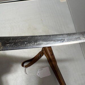 May include: A metal knife blade with the word Finland and the word Stainless engraved on the surface. The blade is positioned above a brown leather sheath that features a small metal snap button and a hanging loop, with a white price tag attached.