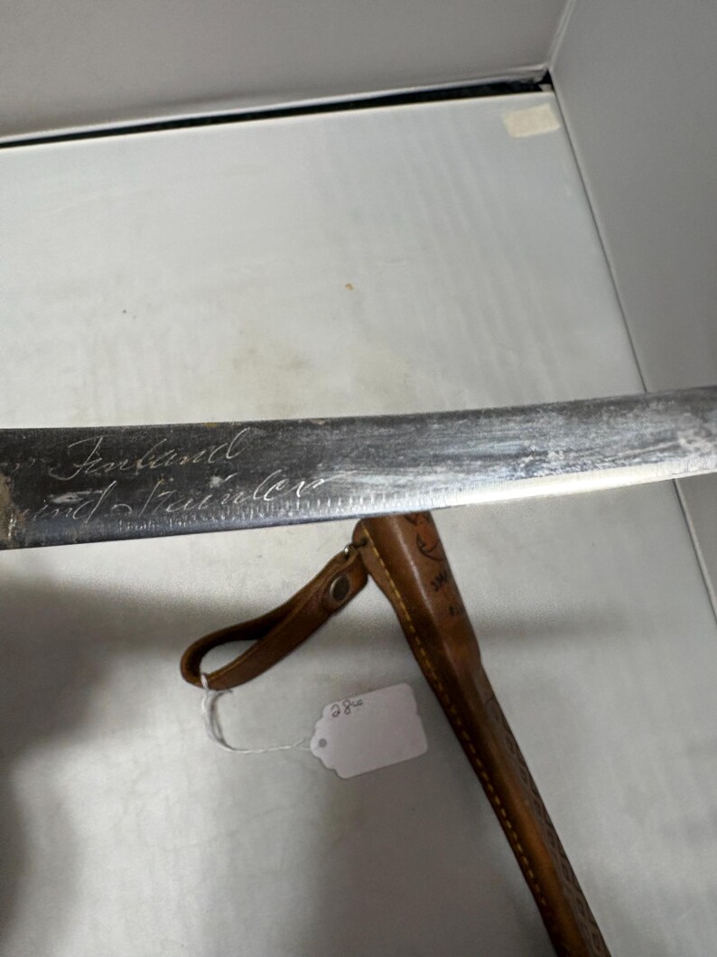 May include: A metal knife blade with the word Finland and the word Stainless engraved on the surface. The blade is positioned above a brown leather sheath that features a small metal snap button and a hanging loop, with a white price tag attached.