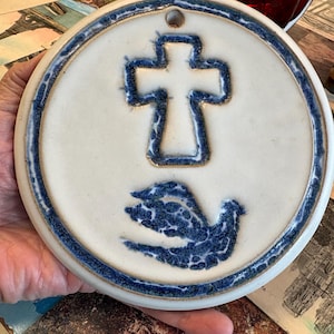 Könnte beinhalten: Eine runde Keramik-Wandplatte mit cremefarbener Basis und blau gesprenkeltem Rand. In der Mitte befindet sich ein erhabenes Kreuzsymbol über einer stilisierten, blau gesprenkelten Taube. Ein kleines Loch oben dient zum Aufhängen.