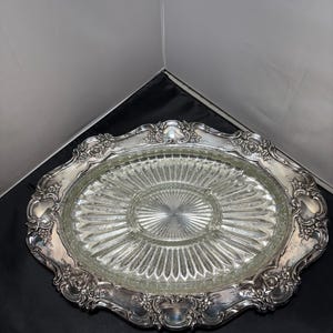 Op de afbeelding: Zilveren serveerschaal met een heldere glazen inzet. De schaal heeft een ovale vorm met een decoratieve, sierlijke rand. De glazen inzet is verdeeld in secties, met een centraal cirkelvormig gebied en uitstralende lijnen.