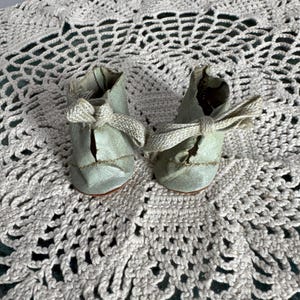 Puede incluir: Un par de zapatos de bebé antiguos en un suave verde apagado. Cada zapato presenta un delicado lazo y costuras intrincadas. Los zapatos se exhiben sobre un tapete de ganchillo color crema, mostrando una estética vintage.