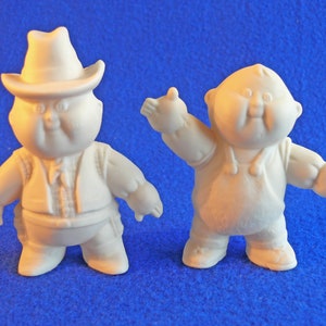 May include: Two white ceramic figurines of chubby characters. One is wearing a cowboy hat and vest. The other is waving with a big smile.