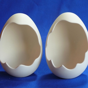 May include: Two white ceramic egg-shaped containers with the top portion removed, revealing the hollow interior. The containers are smooth and have a slightly textured surface.