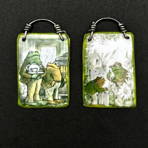 May include: Two rectangular pendants with wire loops. Each pendant features a whimsical illustration of anthropomorphic frogs. One shows frogs indoors, the other outdoors. The pendants have a green border and a silver wire loop.
