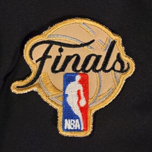 May include: An embroidered patch featuring a tan basketball graphic with the word Finals in black script lettering. A vertical rectangular NBA logo with a white silhouette of a player on a blue and red background is centered over the basketball.